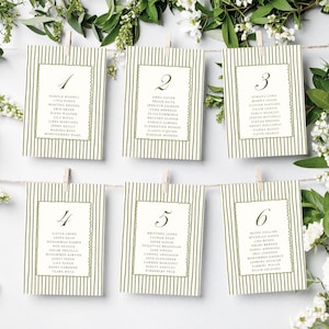 May include: Six ivory place cards with olive green striped backgrounds and scalloped borders are suspended from a string with clothespins. Each card features a number and a list of names. White flowers and greenery frame the cards.