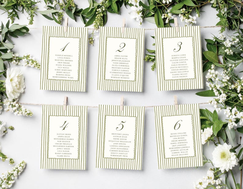 May include: Six ivory place cards with olive green striped backgrounds and scalloped borders are suspended from a string with clothespins. Each card features a number and a list of names. White flowers and greenery frame the cards.