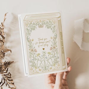 Op de afbeelding: Een wenskaart met een boekontwerp, bloemenillustraties en de tekst "Thank you for being a part of our story." De ruggengraat luidt "Happily Ever After." De kaart wordt door een hand vastgehouden, met een varen en lint op de achtergrond.