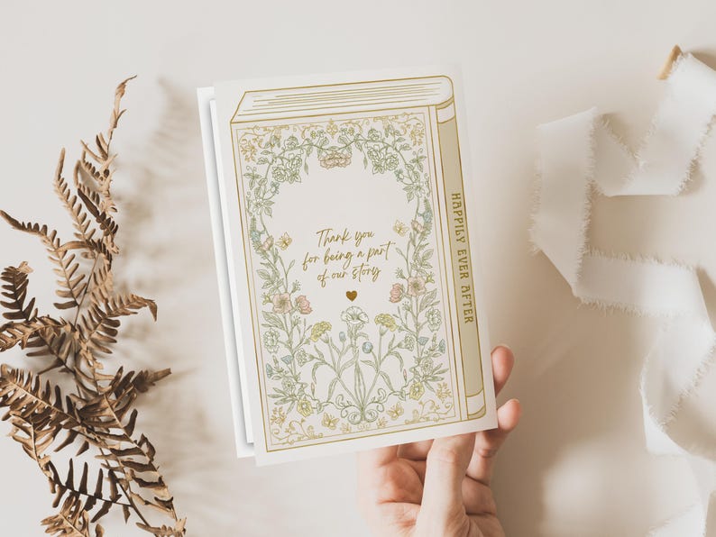 Op de afbeelding: Een wenskaart met een boekontwerp, bloemenillustraties en de tekst "Thank you for being a part of our story." De ruggengraat luidt "Happily Ever After." De kaart wordt door een hand vastgehouden, met een varen en lint op de achtergrond.