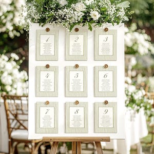 May include: A white seating chart with nine numbered cards, each with a green striped border and gold accents. The chart is adorned with white flowers and greenery, perfect for a wedding or event. The cards display guest names.