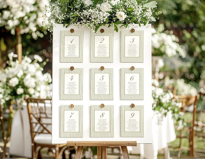 May include: A white seating chart with nine numbered cards, each with a green striped border and gold accents. The chart is adorned with white flowers and greenery, perfect for a wedding or event. The cards display guest names.