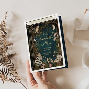 May include: A wedding invitation in the shape of a book with a dark blue background and gold lettering. The text reads "You're Invited Annabelle and Matthew are getting married limited edition volume one". The cover features a whimsical illustration of a forest scene with owls, flowers, and a snake.