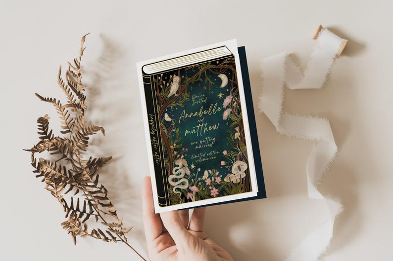 May include: A wedding invitation in the shape of a book with a dark blue background and gold lettering. The text reads "You're Invited Annabelle and Matthew are getting married limited edition volume one". The cover features a whimsical illustration of a forest scene with owls, flowers, and a snake.
