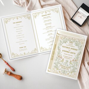 Op de afbeelding: Bruiloft briefpapier set met een programma, uitnodiging en ringdoos. De uitnodiging luidt "Welkom bij de bruiloft van Victoria Warner en Stephen Smith". Gouden bloemmotieven en tekst op een witte achtergrond.