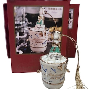 May include: A ceramic champagne bottle ornament in a bucket of ice. The bottle has a green label and a gold bow. The bucket is white with gold accents and the words "Celebrate 2000". A gold string and tassel are attached.