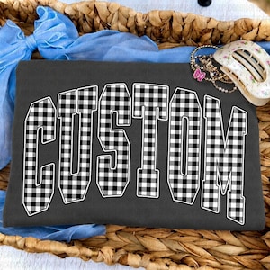 May include: A dark gray t-shirt featuring the word CUSTOM in a black and white buffalo plaid pattern. The shirt is accented with a light blue fabric bow and a white hair clip with a floral pattern, all resting within a woven wicker basket.