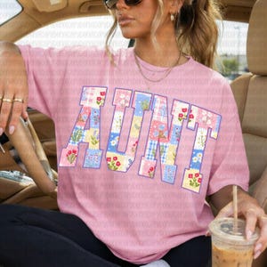 May include: A light pink short-sleeved t-shirt featuring the word AUNT in large block letters. The letters are filled with a patchwork pattern of various floral designs, blue and white gingham checks, and small yellow flowers on a white background.