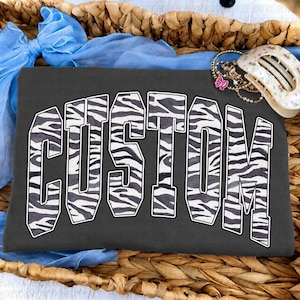May include: A dark gray t-shirt featuring the word CUSTOM in large, block-style letters with a black and white zebra print pattern. The shirt is displayed with a light blue fabric bow and a white hair clip with a floral pattern.