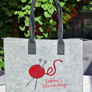Könnte beinhalten: Graue Filztasche mit schwarzen Henkeln und roter Stickerei eines Wollknäuels, Stricknadeln und dem Text "Sabine's Strickzeug".
