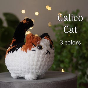 Könnte beinhalten: Ein gehäkeltes Calico-Katzenspielzeug mit schwarzem, orangem und weißem Fell. Die Katze sitzt auf einer grauen Oberfläche.