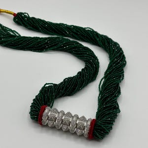 May include: A long green beaded necklace with a silver and white decorative clasp. The necklace is made of many strands of beads and has a red accent on the clasp.