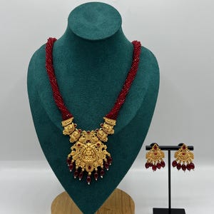 May include: A gold necklace and earring set displayed on a green bust. The necklace has a large, detailed pendant with red bead accents. The earrings echo the pendant's design, also featuring red beads. The necklace is approximately 40 cm long.