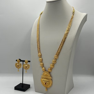 May include: A gold-colored necklace and matching earrings set. The necklace features a long chain with decorative elements and a pendant. The earrings are round with a red stone in the center. The jewelry is displayed on a white mannequin.