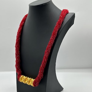 May include: A red beaded necklace with a gold pendant. The necklace is displayed on a black mannequin.