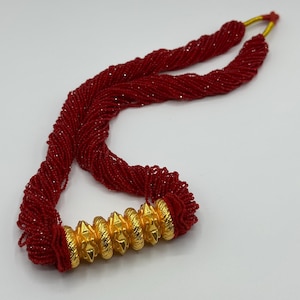May include: A red beaded necklace with a gold pendant. The pendant is made of multiple round pieces with a pointed design.