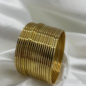 May include: A stack of 20 gold-colored metal bangles. The bangles are all the same size and have a smooth, shiny finish.