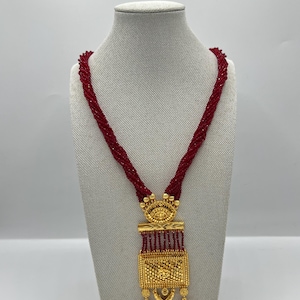 Könnte beinhalten: Eine lange, rote Perlenkette mit einem goldenen Anhänger. Der Anhänger ist rechteckig mit filigranen Details und einem hängenden Design.