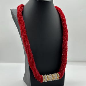 May include: A red beaded necklace with a gold and clear crystal clasp. The necklace is displayed on a black mannequin.