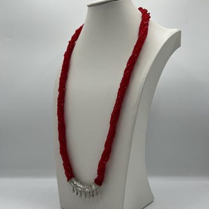 Puede incluir: Un collar largo de cuentas rojas expuesto en un maniquí blanco. El collar está hecho de muchos hilos de pequeñas cuentas rojas, retorcidas juntas. El cierre es de color plateado con dijes colgantes en forma de lágrima.