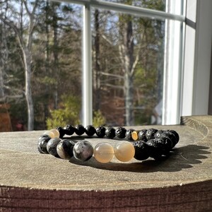 May include: A bracelet made of black lava rock, grey speckled, and light-coloured round beads. The bracelet is displayed on a wooden surface with a window and trees in the background.