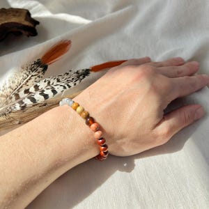 May include: A beaded bracelet featuring a gradient of spherical beads that transition in color from a pale translucent grey to shades of yellow, orange, and deep reddish-brown, arranged in a single strand on an elastic cord.