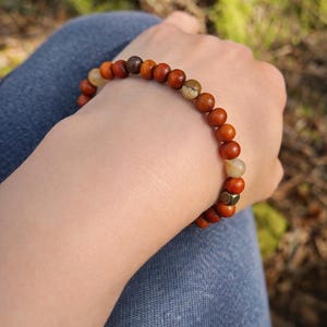May include: A wooden bead bracelet with various shades of brown and tan beads. The bracelet is worn on a wrist, with the background blurred, suggesting an outdoor setting. The beads appear to be approximately 0.5 inches in diameter.