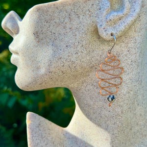 May include: A copper wire earring featuring a series of horizontal loops arranged in a vertical, tapered shape. A small, round, speckled dalmatian jasper bead is attached to the bottom of the wire structure, which hangs from a silver-toned hook.