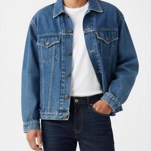 May include: A medium-wash blue denim jacket with button closures and chest pockets. The jacket is worn over a white t-shirt and paired with dark blue jeans. The outfit is casual and classic.