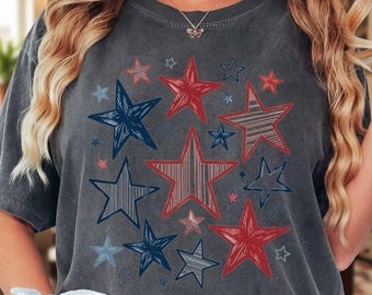 May include: A charcoal gray short-sleeve t-shirt featuring a graphic print of multiple stars in various sizes and styles. The stars are colored in shades of red, white, and blue, with some featuring striped or sketched textures across the front.