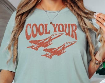 May include: A light sage green short-sleeve t-shirt featuring the text COOL YOUR in a distressed orange font above a graphic illustration of two fighter jets flying in formation. The shirt has a crew neckline and a relaxed fit.