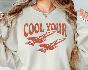 May include: A beige crewneck sweatshirt featuring the text COOL YOUR in a distressed red font, positioned above a graphic illustration of two fighter jets flying in formation. The design has a weathered, vintage-inspired texture.