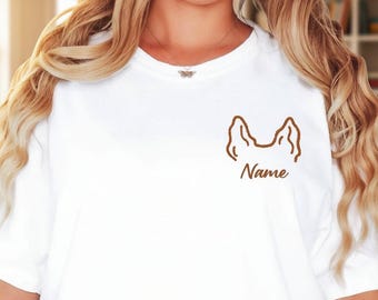 May include: A white t-shirt featuring a brown line art graphic of dog ears on the left chest area. Below the ears, the word Name is printed in a brown cursive script. The shirt has short sleeves and a crew neckline, worn with blue jeans.