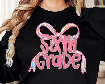 May include: A black long-sleeve sweatshirt featuring a graphic print of a large, iridescent-colored bow with the text "Sixth Grade" written in bold, pink, bubble-style lettering centered directly underneath the bow design on the front.