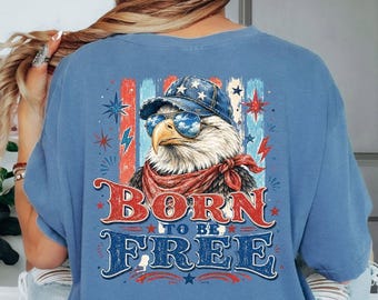 May include: A blue t-shirt featuring a graphic on the back of a bald eagle wearing a blue denim cap and sunglasses, with a red bandana around its neck. The design includes red and blue vertical stripes, stars, lightning bolts, and the text BORN TO BE FREE.