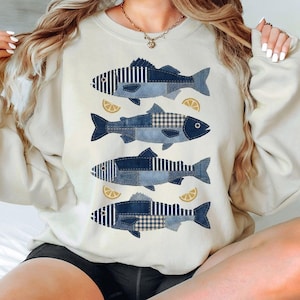 May include: Cream-colored sweatshirt featuring four fish designs in shades of blue denim patchwork. Each fish is accompanied by a lemon slice. The sweatshirt has a relaxed fit and long sleeves.