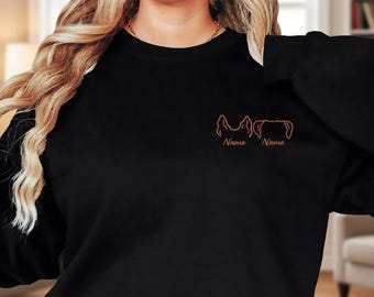 May include: A black long-sleeve sweatshirt featuring an embroidered design on the upper left chest. The design consists of two brown line-art dog ear outlines, each positioned above the word Name in a matching brown cursive script font.