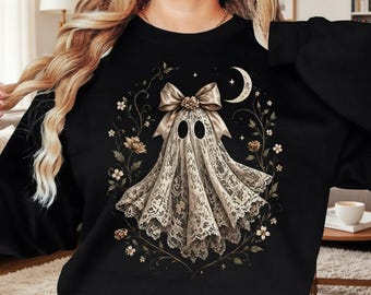 May include: A black long-sleeve sweatshirt featuring a central graphic of a ghost draped in beige lace fabric. The ghost is adorned with a large beige bow at the top and is surrounded by a circular arrangement of delicate floral vines and a crescent moon.