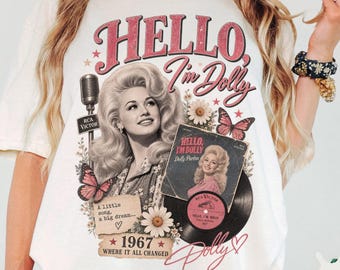 May include: An ivory t-shirt featuring a graphic print of Dolly Parton with the text Hello, I'm Dolly. The design includes a vintage microphone, a vinyl record, butterflies, daisies, and the text 1967 Where it all changed and A little song, a big dream.
