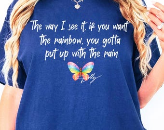 May include: A navy blue short-sleeve t-shirt featuring white text that reads, The way I see it, if you want the rainbow, you gotta put up with the rain. Below the text is a colorful rainbow butterfly graphic and the signature Dolly in white.