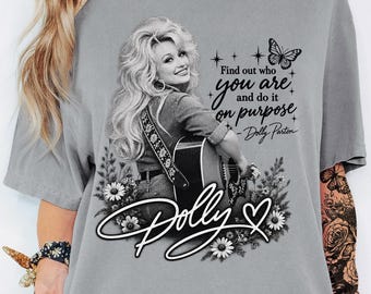 May include: A grey short-sleeved t-shirt featuring a black and white graphic of Dolly Parton playing a guitar. The design includes the text 'Find out who you are and do it on purpose - Dolly Parton', a butterfly, flowers, and the name 'Dolly'.