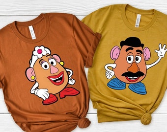 May include: Two brown t-shirts with a cartoon image of Mr. and Mrs. Potato Head. The shirt on the left has Mrs. Potato Head with a pink flower on her head. The shirt on the right has Mr. Potato Head with a mustache and a black hair.