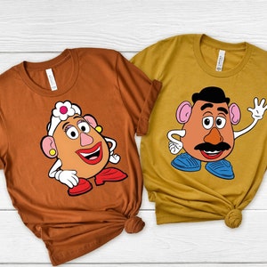 May include: Two brown t-shirts with a cartoon image of Mr. and Mrs. Potato Head. The shirt on the left has Mrs. Potato Head with a pink flower on her head. The shirt on the right has Mr. Potato Head with a mustache and a black hair.