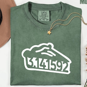 May include: A moss green t-shirt with a white graphic of a pie slice and the numbers 3.141592. The shirt has a crew neck and short sleeves. A gold necklace with a star pendant is draped on the shirt. The shirt is labeled XL-TG-EG.