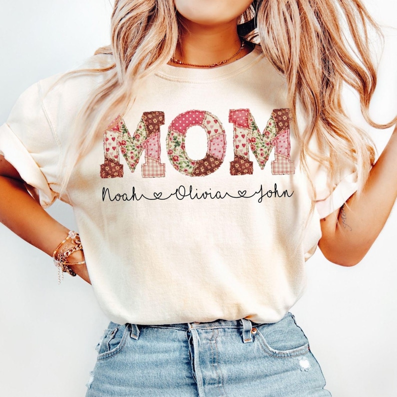 May include: Cream-colored t-shirt with the word "MOM" in a patchwork design of floral and polka dot patterns. Below the word "MOM" are the names "Noah", "Olivia", and "John" with heart symbols.