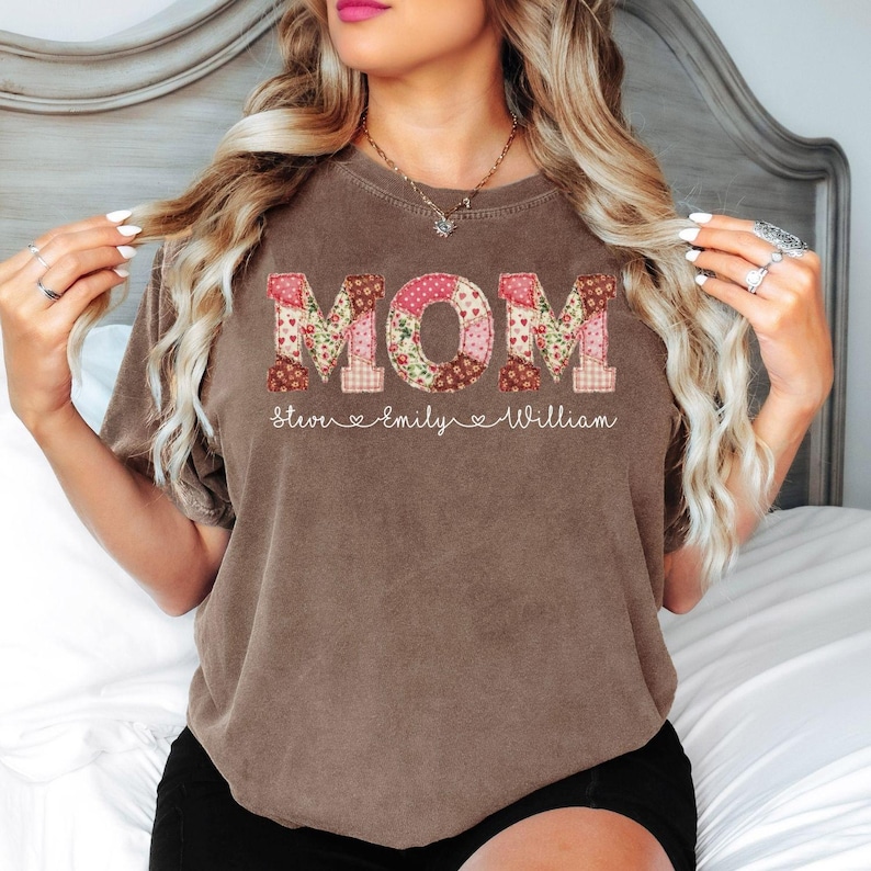 A woman with long blonde hair wearing a brown t-shirt with the word "MOM" printed on it.