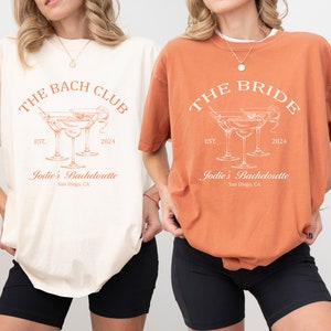 May include: Two t-shirts with a bachelorette party theme. The ivory t-shirt says "The Bach Club" with a cocktail glass graphic and the text "Jodie's Bachelorette, San Diego, CA". The orange t-shirt says "The Bride" with a cocktail glass graphic and the text "Jodie's Bachelorette, San Diego, CA".