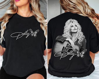 May include: A black t-shirt featuring a white script logo that reads Dolly next to a butterfly graphic on the front. The back displays a large black and white photographic print of a woman with long blonde hair holding a guitar, with a butterfly.