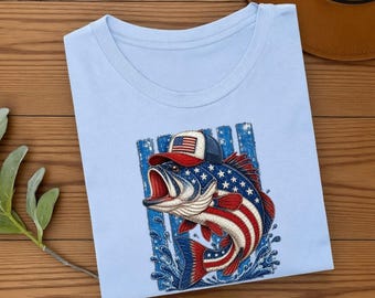 May include: A light blue t-shirt featuring a graphic of a fish wearing a baseball cap. The fish and cap are decorated with a red, white, and blue American flag pattern, including stars and stripes, set against a blue vertical striped background.