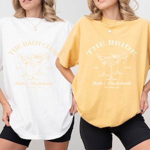 May include: Two oversized t-shirts with a bachelorette party theme. The white t-shirt says "The Bach Club" with a martini glass graphic and the text "Jodie's Bachelorette San Diego, CA". The yellow t-shirt says "The Bride" with a martini glass graphic and the text "Jodie's Bachelorette San Diego, CA".